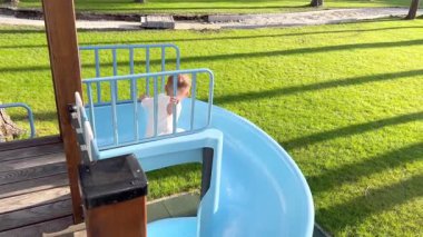 Küçük bir çocuk topun peşinden mavi bir sarmal kaydırağına gidiyor. Bir çocuk çam ormanının yakınındaki bir parkta oyun oynuyor.