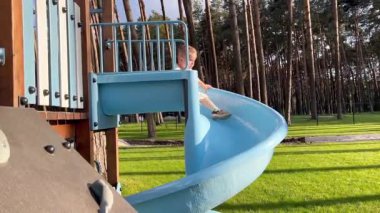 Küçük bir çocuk topun peşinden mavi bir sarmal kaydırağına gidiyor. Bir çocuk çam ormanının yakınındaki bir parkta oyun oynuyor.