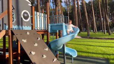 Küçük bir çocuk topun peşinden mavi bir sarmal kaydırağına gidiyor. Bir çocuk çam ormanının yakınındaki bir parkta oyun oynuyor.