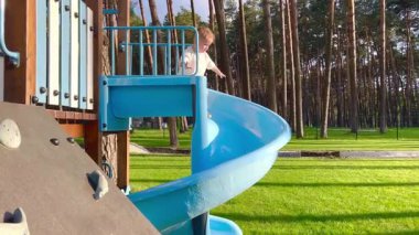 Küçük bir çocuk topun peşinden mavi bir sarmal kaydırağına gidiyor. Bir çocuk çam ormanının yakınındaki bir parkta oyun oynuyor.