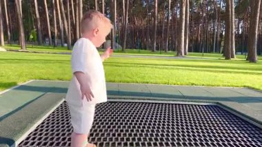  İki yaşında küçük bir çocuk atlıyor, eğleniyor, trambolinde sarkmış metal bir ağ üzerinde ormanın yakınındaki bir parkta.