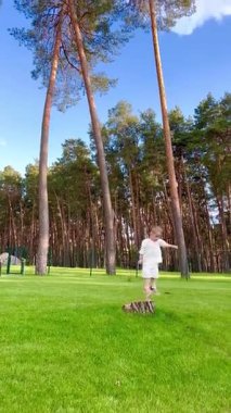 Küçük bir çocuk parkta oynarken eğleniyor, çocuk bir kütüğün üzerinde duruyor ve üzerinden atlıyor.