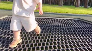  İki yaşında küçük bir çocuk atlıyor, eğleniyor, trambolinde sarkmış metal bir ağ üzerinde ormanın yakınındaki bir parkta.