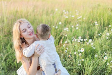 Anne, çayırda papatyaları olan küçük bir çocuğa sarılıyor, tatlı bir anne ve iki yaşındaki bir çocuk yaz tarlasında eğleniyor.