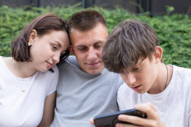 Parkta bir bankta oturan üç genç, akıllı telefonlar kullanarak dışarıda dinlenirken sosyal medyaya göz atıyorlar. Yüksek kalite fotoğraf