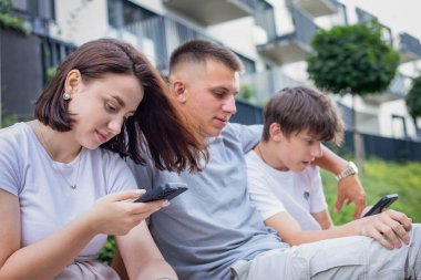 Parkta bir bankta oturan üç genç, akıllı telefonlar kullanarak dışarıda dinlenirken sosyal medyaya göz atıyorlar. Yüksek kalite fotoğraf
