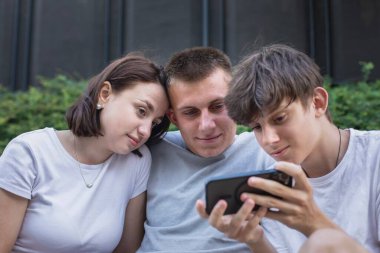 Parkta bir bankta oturan üç genç, akıllı telefonlar kullanarak dışarıda dinlenirken sosyal medyaya göz atıyorlar. Yüksek kalite fotoğraf