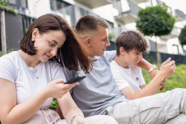 Parkta bir bankta oturan üç genç, akıllı telefonlar kullanarak dışarıda dinlenirken sosyal medyaya göz atıyorlar. Yüksek kalite fotoğraf