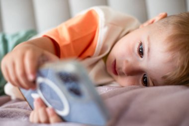 Çocuk yatakta uzanıyor ve büyüleyici bir şekilde akıllı telefon izliyor, modern yaşam tarzını ve teknolojiyi evde nasıl kullandığını gösteriyor. Yüksek kalite fotoğraf