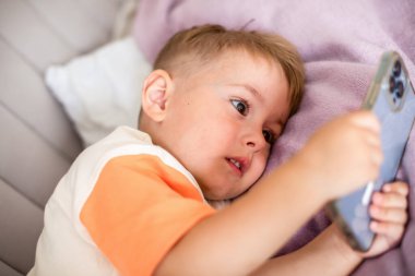 Genç bir çocuk uzanıp akıllı bir telefonla çizgi film izliyor, modern çocukluğu ve evdeki dijital eğlenceyi resmediyor. Yüksek kalite fotoğraf