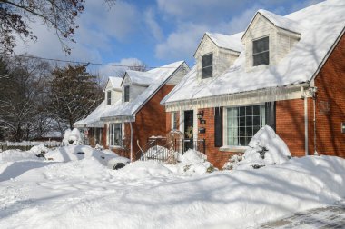 Toronto, ON, Kanada - 27 Şubat 2025: Banliyö evi çatıdan sarkan buz saçaklarıyla kaplı, güneş ışığı beyaz kış manzarasına gölge düşürürken,.