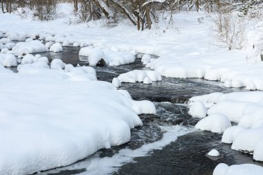 Küçük bir dere karlı bir ormanın içinden akar, karla kaplı kayalar ve kıyılar huzurlu bir kış manzarası yaratır..