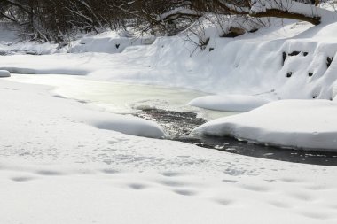 Küçük bir dere karlı bir ormanın içinden akar, karla kaplı kayalar ve kıyılar huzurlu bir kış manzarası yaratır..