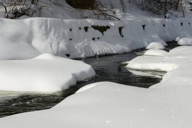 Küçük bir dere karlı bir ormanın içinden akar, karla kaplı kayalar ve kıyılar huzurlu bir kış manzarası yaratır..