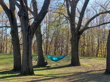 Toronto, ON, Kanada 2 Mayıs 2024: Yüksek Park 'ın sessiz bir köşesinde bir bisiklet dinlenirken, uzun yapraksız ağaçların arasında huzur içinde asılı duran mavi bir hamak...