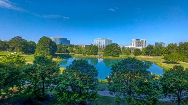 Charlotte, NC, ABD - 28 Nisan 2025: Berrak bir gökyüzü altında modern ofis ve yerleşim binalarıyla çevrili yemyeşil ağaçlar ve küçük bir gölet.