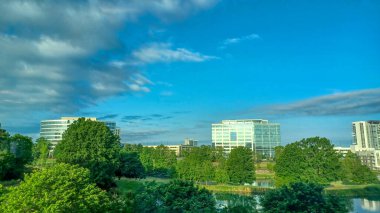 Charlotte, NC, ABD - 28 Nisan 2025: Berrak bir gökyüzü altında modern ofis ve yerleşim binalarıyla çevrili yemyeşil ağaçlar ve küçük bir gölet.