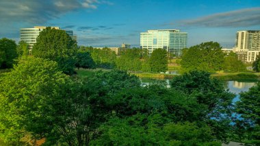 Charlotte, NC, ABD - 28 Nisan 2025: Berrak bir gökyüzü altında modern ofis ve yerleşim binalarıyla çevrili yemyeşil ağaçlar ve küçük bir gölet.