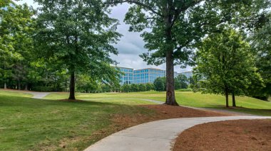 Charlotte, NC, ABD - 30 Nisan 2025: Modern ofis binaları yeşil çimenlerin, ağaçların ve Ballantyne Corporate Park 'ta bulutlu bir gökyüzünün altında bir yürüyüş yolunun ortasında duruyor.