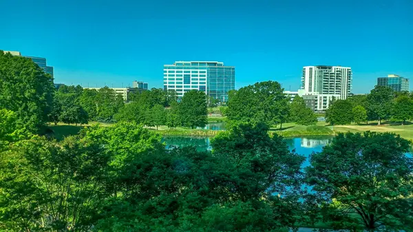 Charlotte, NC, ABD - 28 Nisan 2025: Berrak bir gökyüzü altında modern ofis ve yerleşim binalarıyla çevrili yemyeşil ağaçlar ve küçük bir gölet.