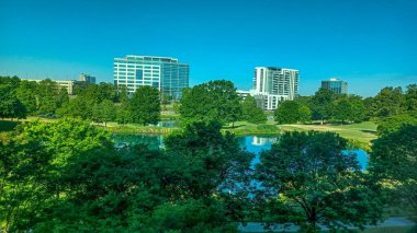 Charlotte, NC, ABD - 28 Nisan 2025: Berrak bir gökyüzü altında modern ofis ve yerleşim binalarıyla çevrili yemyeşil ağaçlar ve küçük bir gölet.