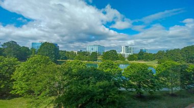 Charlotte, NC, ABD - 28 Nisan 2025: Berrak bir gökyüzü altında modern ofis ve yerleşim binalarıyla çevrili yemyeşil ağaçlar ve küçük bir gölet.