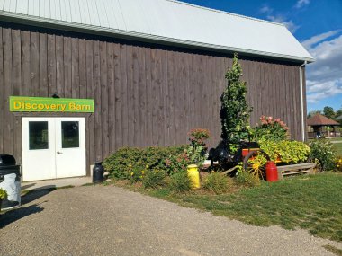 Toronto, ON, Kanada 1 Eylül 2024: Downeys Çiftliği 'nde yeşil çimenler, çam ağaçları ve kabarık bulutlu parlak mavi gökyüzü çerçeveli büyük bir ahırın yanında küçük kahverengi bir ahşap kulübe duruyor...