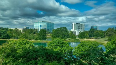 Charlotte, NC, ABD - 28 Nisan 2025: Berrak bir gökyüzü altında modern ofis ve yerleşim binalarıyla çevrili yemyeşil ağaçlar ve küçük bir gölet.