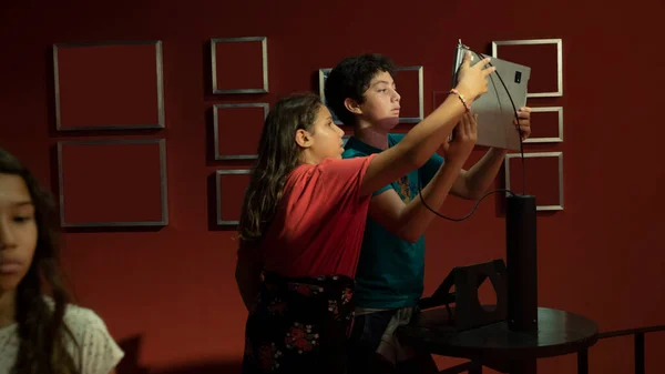 stock image a pre-adolescent boy and girl in an interactive room with red walls holding up a laptop computer, the two children show interest in what they see