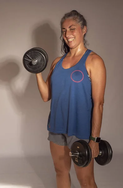 adult woman wearing sports clothes, holding dumbbells and looking at the camera with a smile, is training and sweating after having exercised.