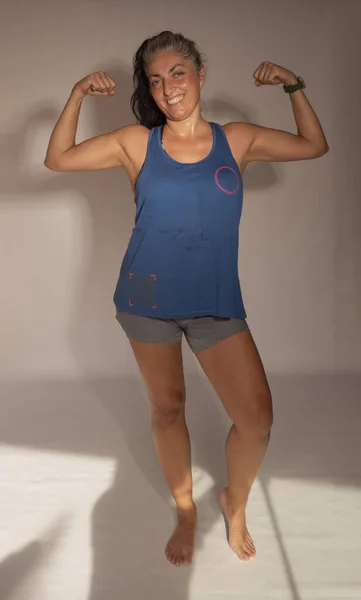 woman around 45 years old with sport clothes, raising her arms showing her biceps, making the gesture of a strong woman. Woman smiling and looking at camera. white studio background. brunette woman with grey hair.