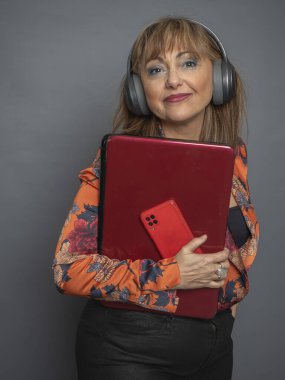 Kameraya bakan girişimci bir kadın, gri arka plan, elinde dizüstü bilgisayar ve cep telefonu, bluetooth kulaklık takıyor..