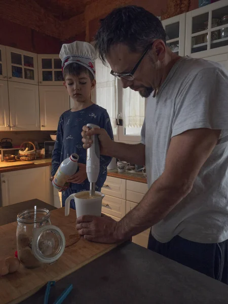 Father and son cooking pancake, the father is with the mixer and the son is with the milk bottle helping his father, they are in a kitchen inside the house, the boy is wearing a chef\'s hat.