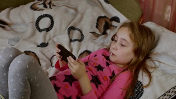 小さな美しい女の子はソファの上にあり スマートフォンの画面を熱心に見て 甘いジューシーなロリポップを吸います 子供や青少年におけるデジタル中毒の問題の概念 — ストック動画
