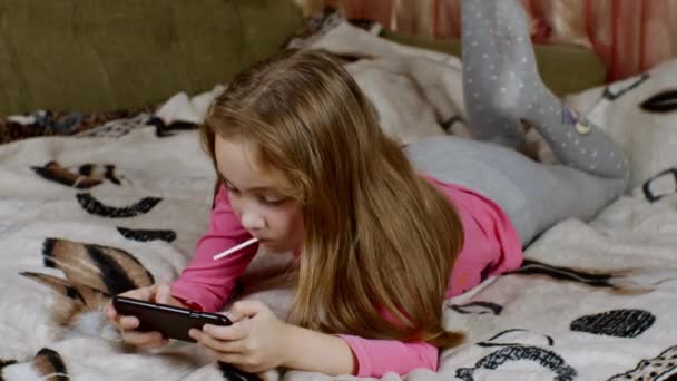Tatlı Sarı Saçlı Ağzında Lolipopu Olan Bacaklarını Sallayan Akıllı Telefon — Stok video