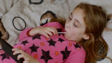 Sol elinde akıllı telefon ve sağ elinde sulu bir lolipop olan bir kız akıllı telefon ekranına dikkatlice bakar ve lolipopu yalar. Sağlık ve modern zımbırtı sorunu kavramı.