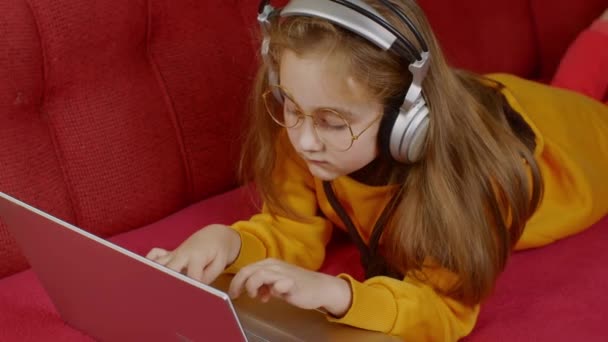 長いブロンドの髪をした面白い女の子は 大きな丸い眼鏡で 黄色のスウェットシャツで ラップトップの前にソファの上にあり コンピュータゲームをプレイし 勝利的に彼女の手をスローします デジタル中毒の概念 — ストック動画