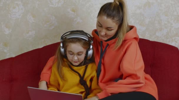 眼鏡とヘッドフォンでかわいい娘を持つ美しい若い母親がソファに座って一緒にノートパソコンを入力しています 子供たちにコンピュータを教えるという概念は — ストック動画