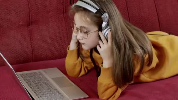 Забавная Маленькая Девочка Наушниках Лежит Диване Смотрит Экран Ноутбука Симпатичная — стоковое видео