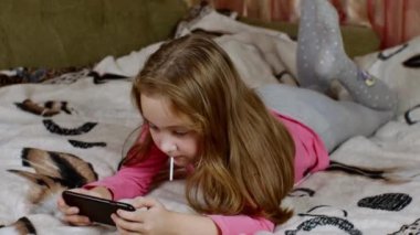 Sevimli sarı saçlı bir kız kanepeye uzanır, bacaklarını sallar, lolipopunu emer, akıllı telefon ekranına bakar ve zafer için elini kaldırır. Çocuk ve ergenlerde dijital bağımlılık kavramı.