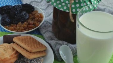 Bardaktaki taze doğal süt, bir kavanoz reçel, beyaz çay kaşığı, kurutulmuş meyvelerle dolu bir çay tabağı ve çok renkli peçetelerin üzerinde kurabiye ve şekerlerle dolu bir fincan tabağı. Süt ürünleri konsepti.