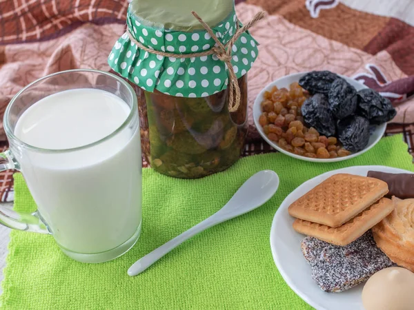 Bardaktaki taze doğal süt, bir kavanoz reçel, beyaz çay kaşığı, kurutulmuş meyvelerle dolu bir çay tabağı ve çok renkli peçetelerin üzerinde kurabiye ve şekerlerle dolu bir fincan tabağı. Süt ürünleri konsepti.