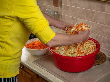 Yakın plan kadın elleri mutfak masasındaki büyük kasede parçalanmış beyaz lahana ve kırmızı havuçları karıştırır. Kış için taze sebzeleri tuzluyorum. Sebze reçelleri.