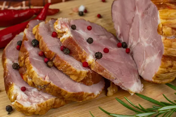 On a wooden cutting board, sliced juicy pork ham, red hot peppers, multi-colored allspice, parsley, dill and basil. Panoramic close-up. The concept of delicious pork products.