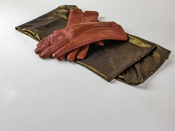Graceful female brown leather gloves lying on a green scarf with a golden tint on a light background. A pair of new brown leather gloves on a green and gold scarf, isolated on a light background.