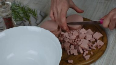 Boiled sausage is cut into small pieces with a kitchen knife on a wooden cutting board for making meat salad. Pepper grinder, sprig of rosemary next to the cutting board. Womens hands close up.