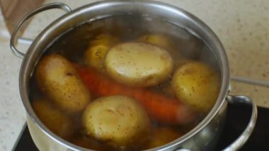 Gas home stove with potatoes and carrots boiled on it in a stainless steel saucepan with a transparent glass lid. Womans hand closes the pot with a lid. The concept of cooking healthy food.