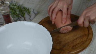 Sausage is cut on wooden cutting board with kitchen knife. Female hands slicing boiled sausage with a knife on a wooden cutting board on a gray kitchen table. Pepper grinder, rosemary sprig. Close-up.