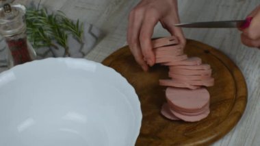 Sausage cut into circles is cut into small pieces on cutting board with knife. Female hands finely chopping sausage with knife on wooden cutting board on gray kitchen table. Preparation of meat salad.