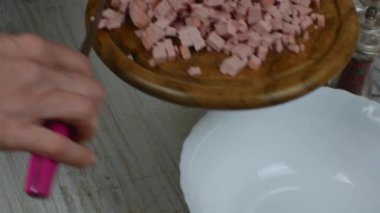 Boiled sausage cut into small pieces on a wooden cutting board is poured into a bowl with a knife. Close-up female hands preparing sausage for meat salad. Housewife prepares meat salad.